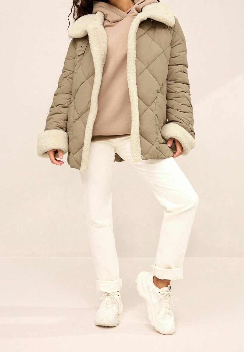 Chaqueta acolchada de color verde oliva con cuello y puños de piel sintética crema, superpuesta a una sudadera beige, combinada con pantalones blancos y zapatillas blancas de suela gruesa.