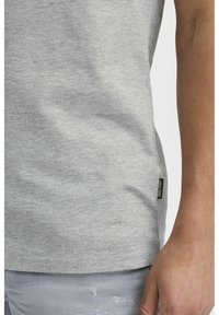 Tricou din bumbac gri cu o textură netedă, mâneci scurte și un mic etichetă neagră la tiv, asortat cu blugi gri deschis, deteriorați.