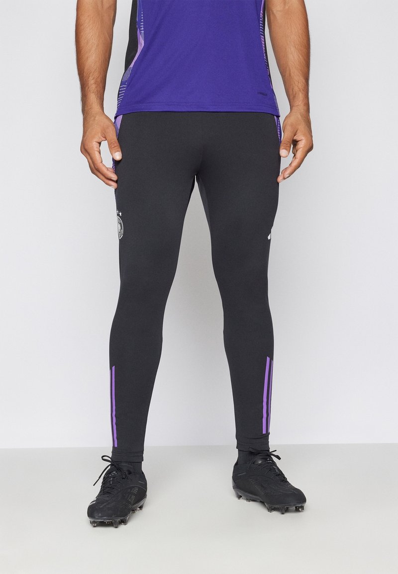 Leggings atletici neri con strisce laterali viola, design aderente, materiale elasticizzato, abbinati a scarpe da calcio nere con tacchetti.