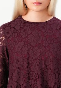 Prim-plan cu o persoană îmbrăcată într-un top din dantelă florală de culoare purpurie închisă, cu mâneci din dantelă transparentă și decolteu rotund.