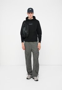 Ung mand iført sort hættetrøje, grå cargo bukser, sneakers, kasket og skuldertaske, stående mod en hvid baggrund.