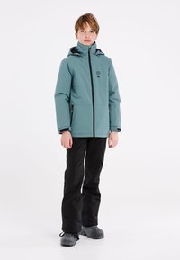 Veste étanche teal avec une fermeture éclair à l'avant et une capuche, bordures noires, logo sur la poitrine, associée à un pantalon noir et des bottes grises.