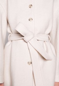 Manteau texturé couleur crème avec boutons en écaille de tortue et ceinture en tissu nouée à la taille.