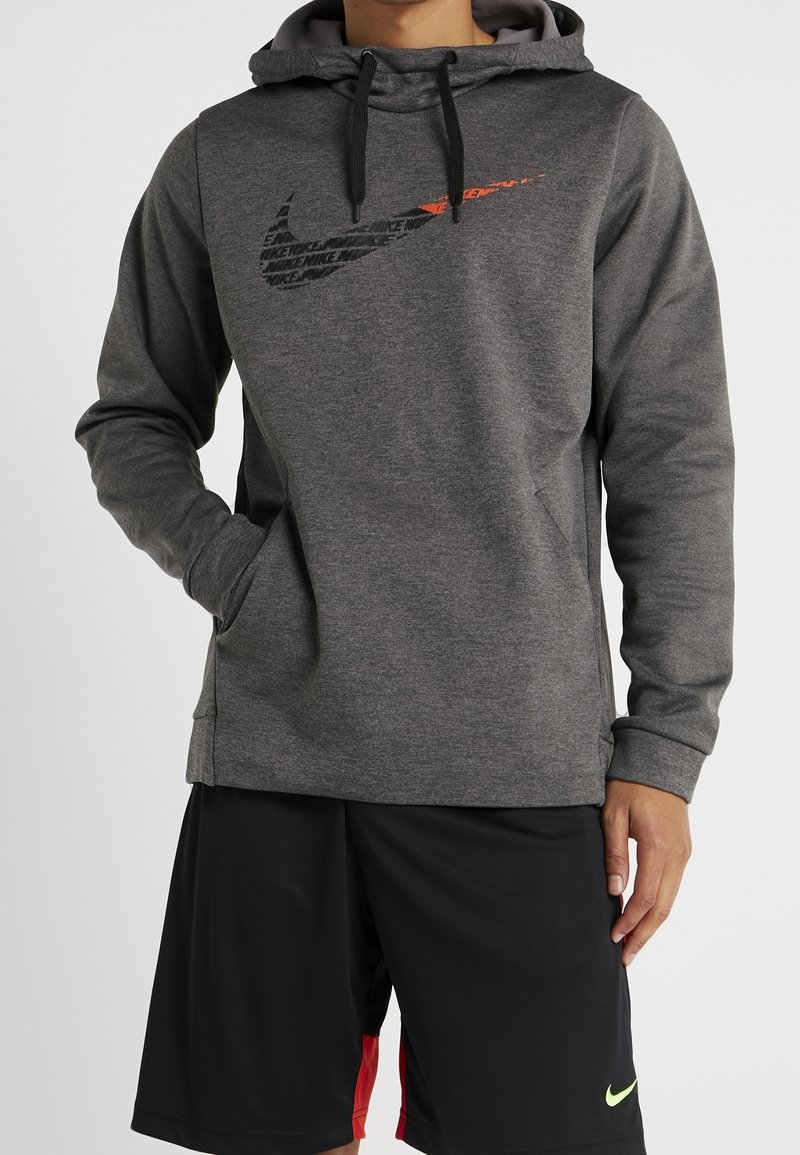 Sweatshirt à capuche gris avec des cordons noirs et un logo Nike réfléchissant orange. Porté sur un short noir avec un accent rouge et un logo vert.