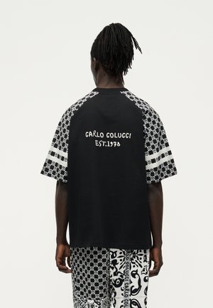 Mężczyzna zwrócony tyłem, ubrany w czarno-białą koszulę w wzory z napisem „Carlo Colucci EST. 1970” oraz dopasowane wzorzyste spodenki.