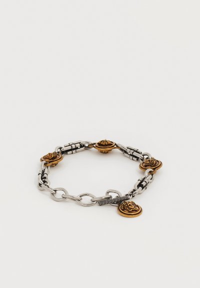 Ασημένιο βραχιόλι αλυσίδας με πολλαπλά χρυσαφένια μενταγιόν που μοιάζουν με νομίσματα και φέρουν ανάγλυφα πρόσωπα, εμφανιζόμενο σε απλό λευκό φόντο.