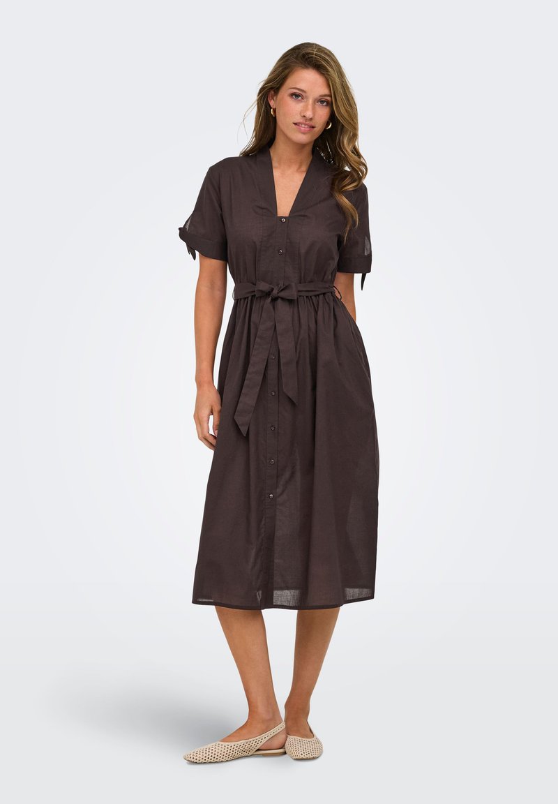 Femme debout portant une robe midi marron foncé boutonnée, à manches courtes et avec une ceinture à la taille, associée à des chaussures plates tissées beiges.