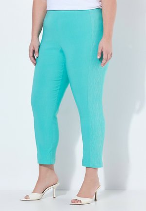 Kvinde iført turkis smalle ankelbukser med subtile lodrette syninger, kombineret med hvide højhælede sandaletter med åben tå.