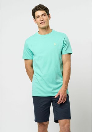 Majica od pamuka mente zelene boje s okruglim izrezom, kratkim rukavima i malim žutim logotipom na prsima. Uparena s tamnoplavim hlačicama.