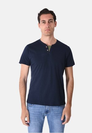 Uomo che indossa una maglietta henley blu navy a maniche corte con due bottoni e jeans azzurri, in piedi davanti a uno sfondo chiaro uniforme.