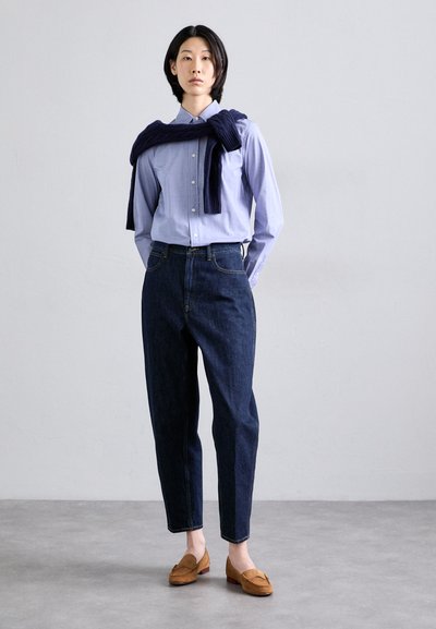 Button-up light blue shirt, dark blue tapered jeans, navy sweater draped over shoulders, and brown loafers on a plain gray background.