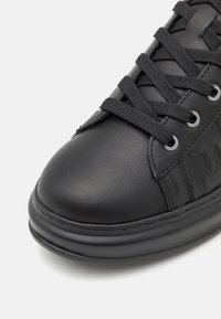 Zwarte sneaker met een glad leren bovenwerk, platte veters, metalen veterogen en een gestructureerd ontwerp aan de zijkant. Ronde neus en dikke zool.