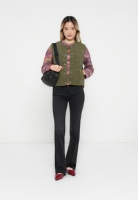 Olijfgroen fleece vest met drukknoopjes, gedragen over een multikleurige gebreide sweater, gecombineerd met zwarte flared jeans en rode schoenen.