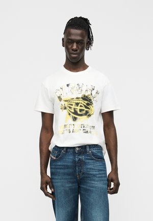 Young man wearing a white graphic T-shirt with a yellow and black frog design and blue jeans, standing against a plain background.