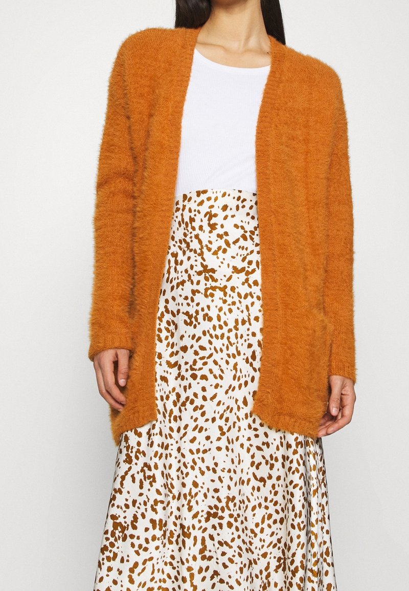 Pomarańczowy, puszysty kardigan z długimi rękawami i otwartym przodem, zestawiony z białą spódnicą w brązowe plamki. Miękka faktura i luźny krój.