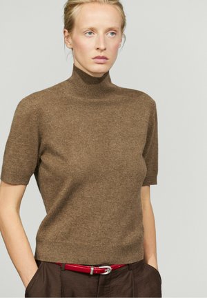 Femme blonde portant un pull à col roulé marron à manches courtes, pantalon marron foncé et ceinture rouge, mains dans les poches, fond neutre.