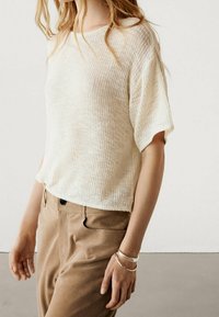 Lys beige tekstureret strikbluse med korte ærmer, parret med tan bukser. Bemærkelsesværdige detaljer inkluderer en afslappet pasform og synlig ribbing.
