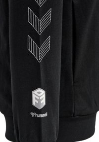 Svart sweatshirt med en texturerad tyg, med vita chevronmönster på ärmarna och en logotyp tryckt nära manschetten.