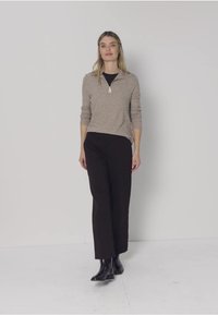 Lichtbruine trui met ritssluiting en lange mouwen, gecombineerd met zwarte wijd uitlopende broek en zwarte enkellaarzen. Eenvoudig ontwerp tegen een neutrale achtergrond.