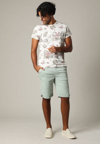 Man met krullend haar, gekleed in een T-shirt met bloemenprint, lichtgroene shorts en witte instappers, staande tegen een effen achtergrond.