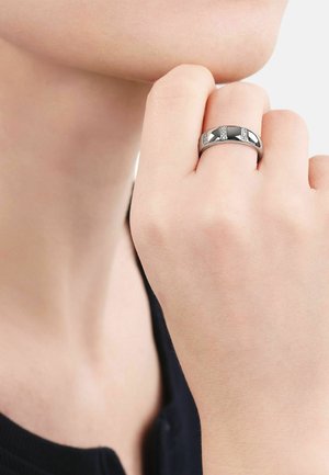 Silberner Ring mit drei vertikalen Reihen kleiner Diamanten, getragen am Finger einer Person nahe dem Kinn vor weißem Hintergrund.