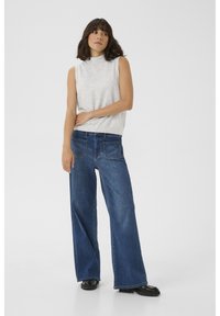 Grijze mouwloze top van gebreide stof, gecombineerd met wijde donkerblauwe denimjeans met achterzakken en gestikte naden. Zwarte schoenen.