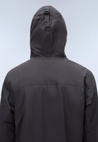Personne portant une veste noire à capuche, vue de dos, montrant un détail de boucle sur la capuche sur un fond clair uni.