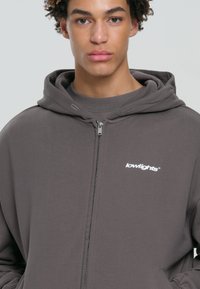 Šedá mikina s kapucňou na zips vyrobená z mäkkého materiálu, s predným zipsom, kengurími vreckami a bielym logom značky na hrudi.