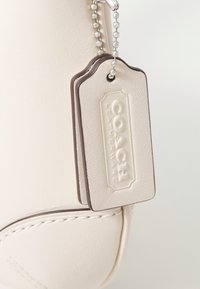 Balta odinė rankinė su pilkai spalvotu žymekliu, pritvirtintu sidabrine grandinėle, su įspaustu užrašu "COACH LEATHERWARE", pasižyminti lygiu paviršiumi ir tvarkingu siuvimu.