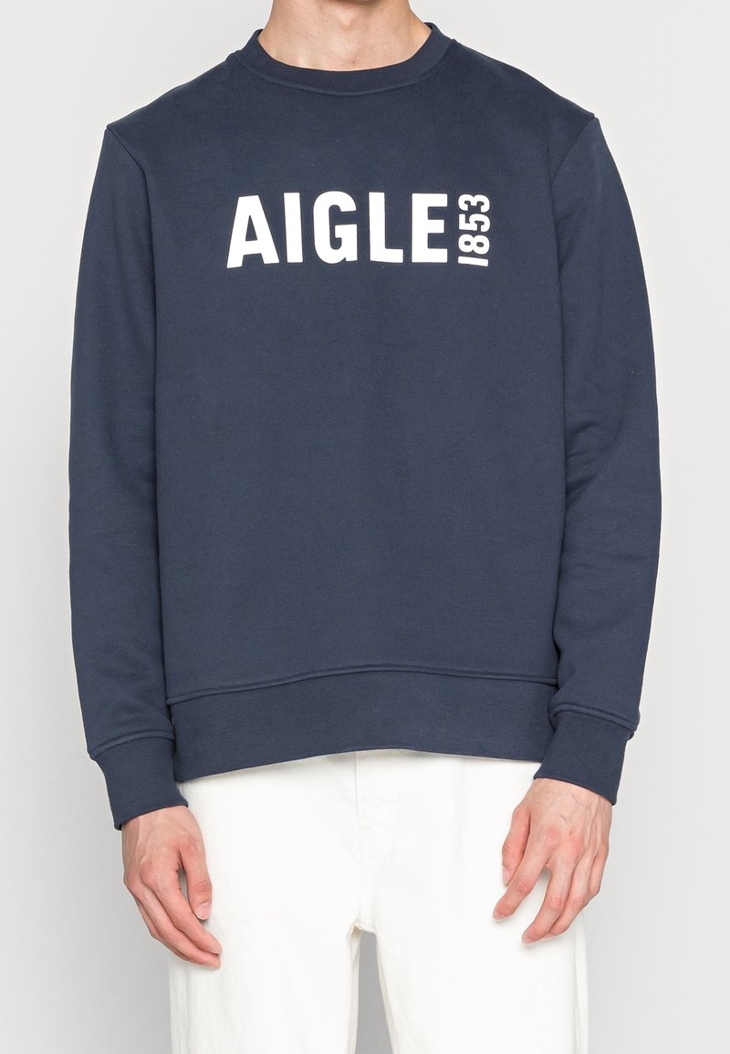 Homme portant un sweat-shirt bleu marine avec le texte blanc "AIGLE 1853" sur la poitrine, associé à un pantalon blanc, debout devant un fond uni.