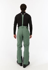 Persoon in groene skibroek met zwarte verstelbare bretels en een zwart lange mouwen shirt, staand met de rug naar de camera op een witte achtergrond.
