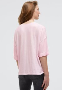 Woman with long brown hair wearing a loose-fitting light pink short-sleeve shirt and dark gray jeans, facing away against a plain background.