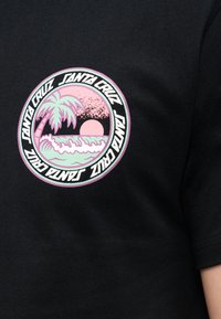 Μαύρο πουκάμισο με κυκλικό παστέλ λογότυπο που απεικονίζει φοίνικα, κυματισμούς του ωκεανού, ηλιοβασίλεμα και το κείμενο "Santa Cruz" επαναλαμβανόμενο γύρω από την άκρη.