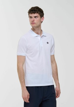 Giovane uomo che indossa una polo bianca semplice con un piccolo logo circolare sul petto, che guarda verso sinistra contro uno sfondo chiaro.