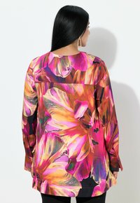 Kvinde med langt sort hår iført en lys blomsterprintet bluse med lange ærmer i pink, orange og lilla nuancer, vendt væk fra kameraet.