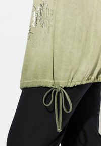 Olivgrünes Baumwolloberteil mit einem gerafften Saum und Kordelzugdetails. Hat auf der Vorderseite bedruckte Akzente in Schwarz und Silber. Kombiniert mit schwarzen Hosen.
