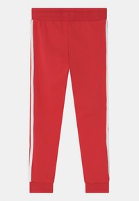 Červené teplákové kalhoty s hladkým povrchem, se bílými bočními pruhy a elastickými manžetami. Pas je také elastický pro pohodlí a dobré padnutí.