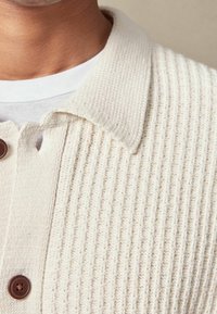 Nahaufnahme einer Person, die eine cremefarbene, gerippte Strickjacke mit Knöpfen über einem weißen Rundhalsausschnitt-Shirt trägt, wobei der Kragen und die Knöpfe sichtbar sind.