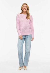 Jeune femme souriante, portant un pull en tricot rose, un jean bleu clair et des talons beiges, debout devant un fond blanc.