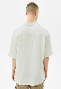 Camicia a maniche corte, di colore chiaro, con colletto, caratterizzata da una texture liscia e una vestibilità comoda, vista da dietro.