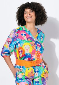 Blusa colorata con un design multicolore a motivi di volti illustrati. Ha una chiusura frontale con bottoni e una cintura arancione a vita arricciata.