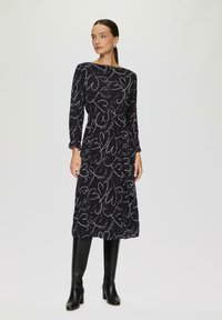 Langes schwarzes Kleid mit weißen Herzen, mit langen Ärmeln und einem Taillengürtel. Kombiniert mit knielangen schwarzen Stiefeln für einen vollständigen Look.