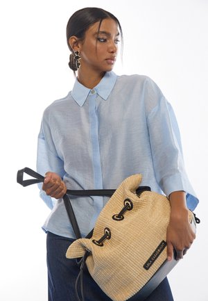 Camisa azul claro de manga corta con botones y corte holgado; sostiene un bolso tejido color beige con correas negras y detalles distintivos en el cierre.