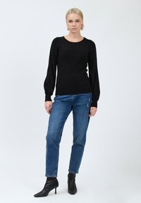 Donna in piedi che indossa un maglione nero a maniche lunghe, jeans blu con leggere abrasioni e stivaletti neri con tacco su uno sfondo bianco uniforme.