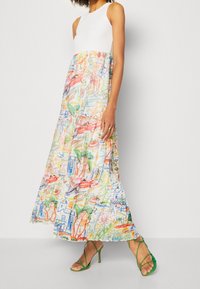 Robe maxi avec un haut blanc, jupe colorée à motifs présentant des designs abstraits, ourlet à volants, et portée avec des talons verts à lanières.