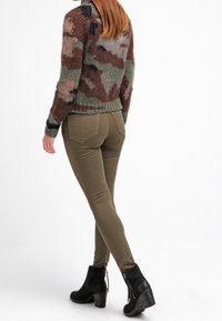 Nainen, jolla on päällään ruskea ja vihreä camouflage-neule, oliivinvihreät kireät housut ja mustat korkeat nilkkasaappaat, kävelee poispäin valkoista taustaa vasten.