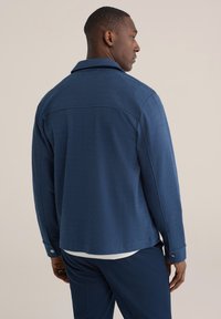 Tumši zila austā jaka ar apkakli, pogas pie atlokiem un vaļēju piegriezumu, redzama no aizmugures uz modeļa.