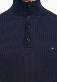 Navyblauwe geribbelde trui met hoge kraag en vier zwarte knopen, featuring een klein rood, wit en blauw logo op de borst.