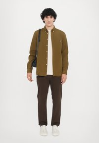 Cămașă maro cu nasturi peste un tricou crem, asortată cu pantaloni maro închis și adidași albi. Prezintă un logo mic și colorat pe cămașă.