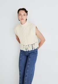 Cream knitted sleeveless sweater with cable pattern and fringe hem, paired with blue high-waisted straight-leg jeans.